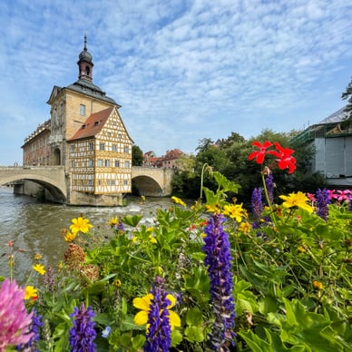 Fachwerkgebäude auf einer Brücke über einen Fluss, umgeben von bunten Blumen.