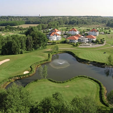 Luftaufnahme eines Golfplatzes mit Teich und Gebäuden im Hintergrund.