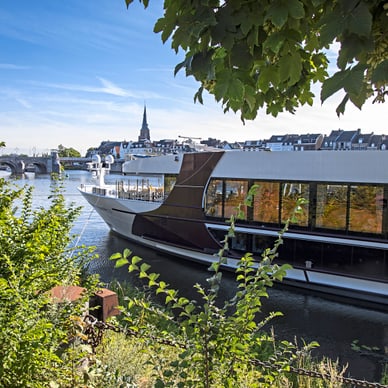 Grosses Boot auf einem Fluss, umgeben von Bäumen und Gebäuden im Hintergrund.