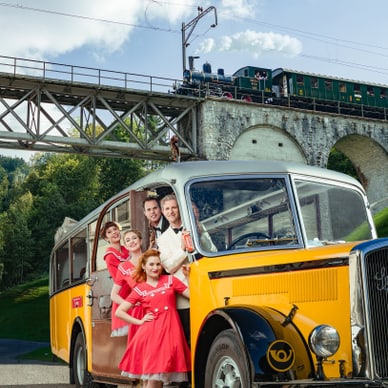 Gruppe in 1950er-Jahre Kleidung vor gelbem Oldtimer-Bus mit Eisenbahnbrücke im Hintergrund.