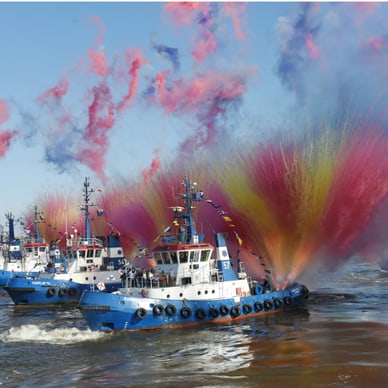 Mehrere Schlepperboote sprühen bunte Wasserfontänen im Hafen.