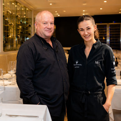Ein Mann und eine Frau in schwarzen Hemden stehen in einem Restaurant mit gedeckten Tischen.