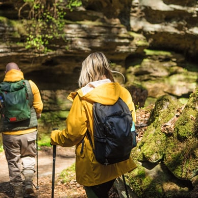 Mann und Frau mit Rucksäcken und Wanderstöcken wandern durch moosbedeckte Felsen im Wald.
