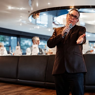Person in Anzug spricht in ein Mikrofon in einem eleganten Restaurant mit Gästen im Hintergrund.