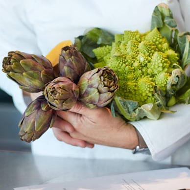 Koch hält Artischocken und Romanesco im Arm neben Rezeptblatt.