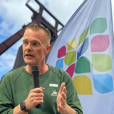 Person hält Mikrofon und spricht neben einer farbenfrohen Flagge und Stahlkonstruktion.