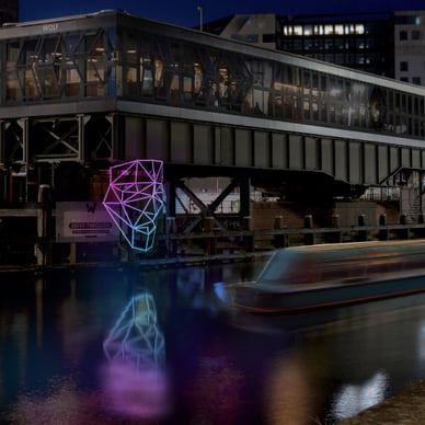 Person mit Mantel steht am Flussufer, Neonlichtkunst und Gebäude im Hintergrund.