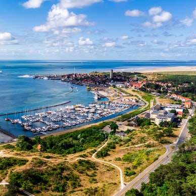 Luftaufnahme des Yachthafens von West-Terschelling mit Leuchtturm, roten Dächern und grünen Hügeln an der Küste.