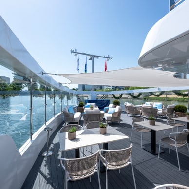 Leeres Sonnendeck der Excellence Crown mit weissen Tischen, Korbstühlen und Blick auf einen Fluss mit Stadtkulisse.