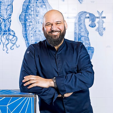 Ein lächelnder Mann in dunklem Hemd steht vor einer blau-weissen Wand mit maritimen Motiven.