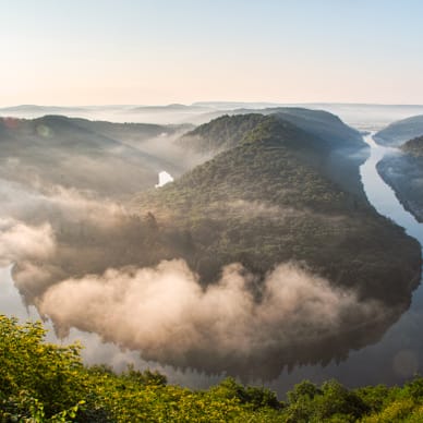 Luftaufnahme einer Flussbiegung mit bewaldeten Hügeln im Nebel.