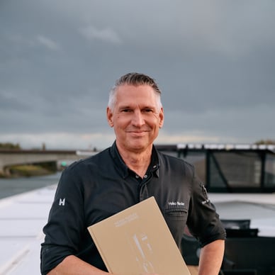 Person mit Kochjacke hält Buch auf einem Boot.