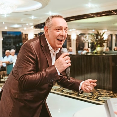 Ein Mann in einem Anzug singt in ein Mikrofon vor einem Publikum in einem eleganten Raum.