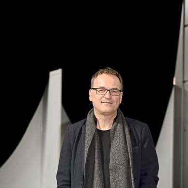 Mann mit Brille und Schal steht vor dunklem Hintergrund.