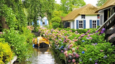 Blumen gesäumter Kanal mit Boot und Strohdachhäusern.