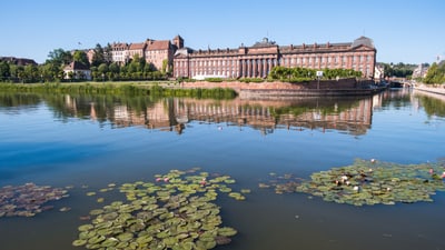 twerenbold_101614.jpg Gebäude am Ufer eines ruhigen Flusses mit Seerosen im Vordergrund.