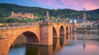 Historische Steinbrücke über einen Fluss vor einer malerischen Altstadt und Hügeln bei Sonnenuntergang.