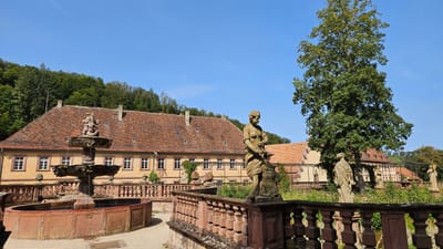 kloster-bronnbach.jpg Historische Gebäude mit Statuen und Bäumen im Vordergrund an einem sonnigen Tag.