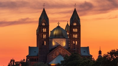 twerenbold_85522.jpg Majestätische Kathedrale bei Sonnenuntergang mit drei Türmen vor orangefarbenem Himmel.