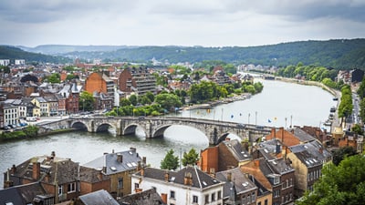 Stadtlandschaft mit Fluss und Brücke, umgeben von Gebäuden und grünen Hügeln.