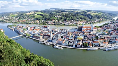 twerenbold_70316.jpg Luftaufnahme einer Stadt an einem Fluss mit Brücken und Booten, umgeben von Hügeln.