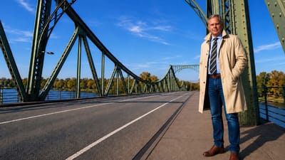 Person in beigem Mantel steht auf einer Brücke mit markanten Metallbögen.