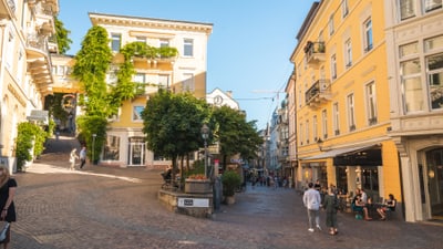 twerenbold_84637.jpg Eine gepflasterte Strasse mit alten Gebäuden, Bäumen und Menschen, die herumlaufen.