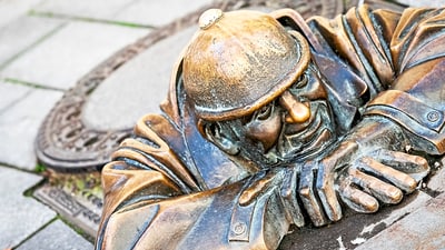 Bronzestatue eines Mannes, der aus einem Kanaldeckel schaut.