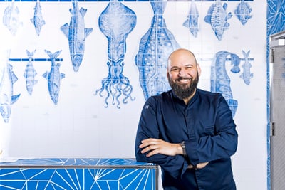 Mann im dunklen Hemd steht vor einer blauen Fliesenwand mit Fischmotiven.