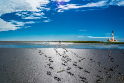 Strandlandschaft mit Fussspuren im Sand, zwei Personen in der Ferne und einem Leuchtturm am Horizont.