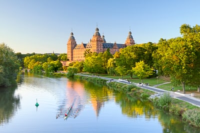 Grosses Schloss am Fluss mit Ruderboot, umgeben von Bäumen.