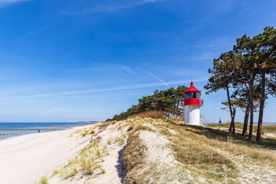 Kleiner Leuchtturm auf sandiger Düne mit Meer im Hintergrund.