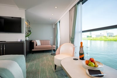 wohlfuehlorte-mit-blick-auf-den-fluss-die-suiten-und-kabinen.jpg Luxuriöses Zimmer mit Sofa, Tisch, Obstschale, Fenster mit Flussblick.