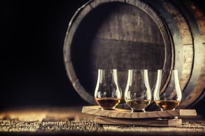 Drei Whiskygläser auf Tablett vor einem Fass.