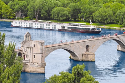 Alte Steinbrücke über Fluss mit Kreuzfahrtschiff und Bäumen im Hintergrund.