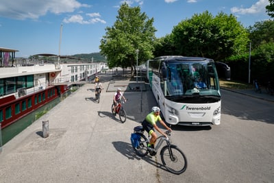 Menschen fahren mit Fahrrädern neben einem geparkten Reisebus und einem Flusskreuzfahrtschiff.