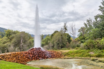 Wasserfontäne, die aus einem Steinhaufen in einer grünen Landschaft aufsteigt.