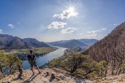wachau.jpg Person auf einem Felsen mit Blick auf einen Fluss und Berge bei Sonnenschein.