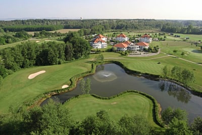 Luftaufnahme eines Golfplatzes mit Teich und Gebäuden im Hintergrund.