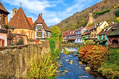 kaysersberg.jpg Eine malerische Altstadt mit Fachwerkhäusern, Fluss und herbstlichen Bäumen.
