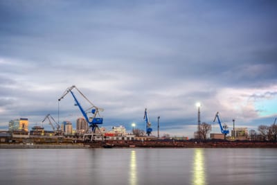 bratislava-aus-einer-anderen-perspektive-auf-der-faehrte-wah.jpg Flussufer mit Kränen und städtischer Skyline im Hintergrund unter bewölktem Himmel.
