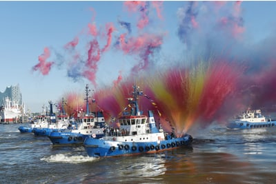 schlepperballett-am-hamburger-hafengeburtstag.jpg Mehrere Schlepperboote sprühen bunte Wasserfontänen im Hafen.