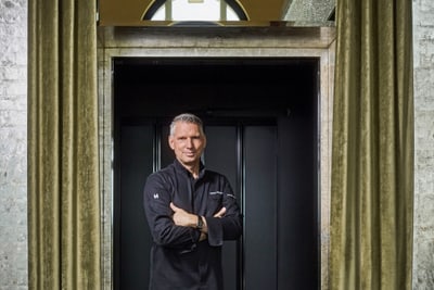 das-excellence-gourmetfestival-10.jpg Mann in schwarzer Kleidung steht mit verschränkten Armen vor Vorhang.
