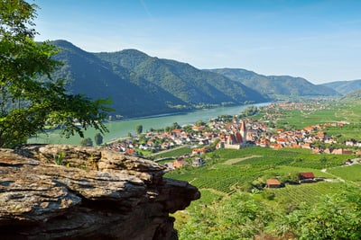 Blick auf ein Tal mit Fluss, Bergen und einer kleinen Stadt mit roten Dächern.