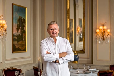 das-excellence-gourmetfestival-10.jpg Mann in weissem Hemd steht in einem eleganten Esszimmer mit Kerzen und Gemälden.