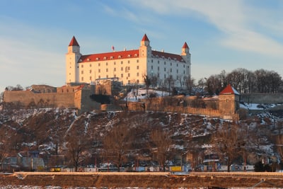 aussicht-auf-die-burg-von-bratislava.jpg Ein Schloss auf einem Hügel mit Blick auf einen Fluss und Spiegelung im Wasser.