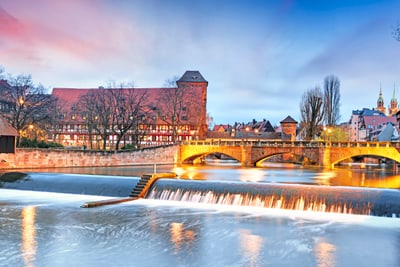 blick-auf-die-maxbruecke-in-nuernberg.jpg Fluss in Nürnberg mit beleuchtetem Brückenbogen und Gebäuden im Hintergrund.