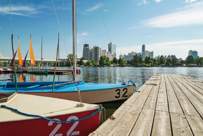 Hölzerner Steg am Wasser mit kleinen Segelbooten und Stadtansicht im Hintergrund.