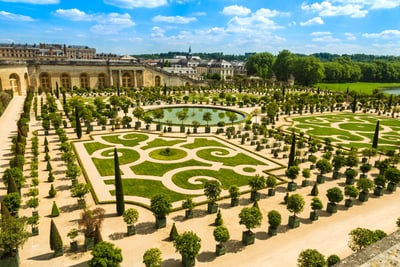 Formaler Garten mit symmetrischen Hecken und Springbrunnen vor einem historischen Gebäude.