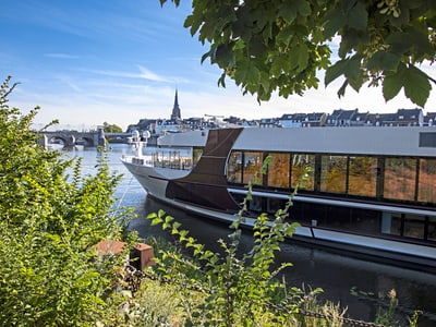 Grosses Boot auf einem Fluss, umgeben von Bäumen und Gebäuden im Hintergrund.